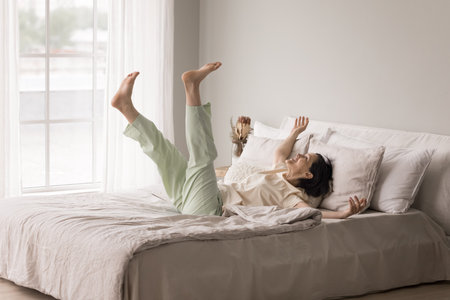Morning luxury comfort. Joyful elderly lady in pajamas throw herself back on big king size bed in light bedroom feel full of energy vitality after good night rest on soft mattress clean natural linenの写真素材