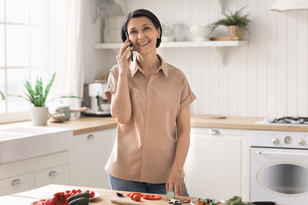 Cooking and chatting. Happy smiling middle aged female retiree speaking on phone hold pleasant conversation distracted from preparing vegetable dish with fresh products discuss diet recipe with friendの写真素材