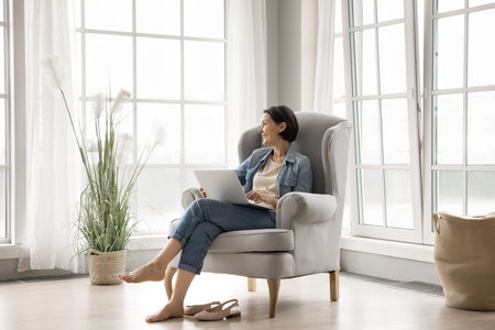 Imagination and computer. Smiling dreamy senior woman relax with notebook in comfortable armchair by panoramic window in living room imagine future plans create ideas visualize distracted from screenの写真素材