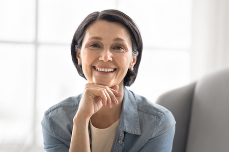 Beautiful aging. Head shot portrait pretty mature female pensioner sit on sofa looking at camera with serene toothy smile touch face express happiness vitality natural beauty of golden years self careの写真素材