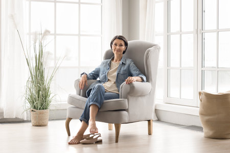 Comfortable home life. Full length portrait happy beautiful older woman retiree relax in comfy armchair look at camera place feet on warm floor enjoy cozy domestic interior in light stylish apartmentの写真素材