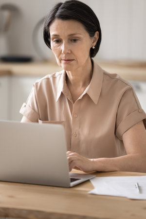 Online research and analysis. Portrait serious older business lady working on freelance from home with digital and paper documents study marketing report analyze data concentrated on notebook screenの写真素材