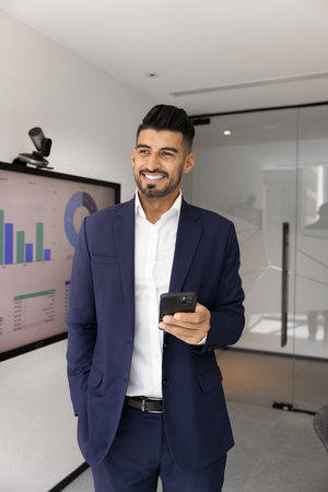 Happy dreamy young Indian marketing expert man holding smartphone, standing at electronic whiteboard with analytic information, sales charts, using mobile phone for messaging. Vertical shotの写真素材
