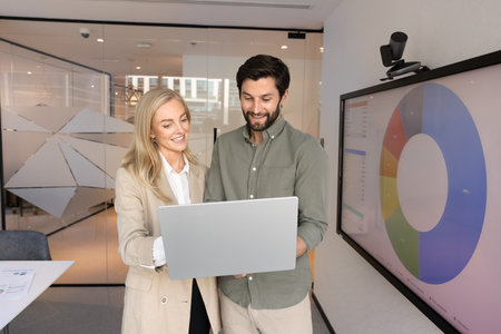 Two positive online product managers preparing marketing reports, analytic information for training presentation, using laptop at electronic whiteboard, analyzing statistic research resultsの写真素材