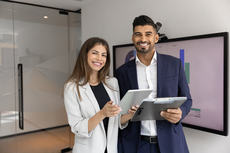 Two cheerful happy multiethnic coworkers analyzing electronic and paper reports, meeting in office boardroom, holding digital tablet and documents, looking at camera, smiling, for portraitの写真素材