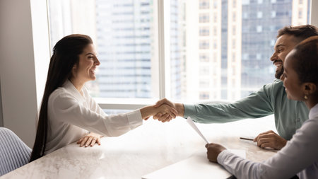 Welcoming new hire. Smiling business team of young diverse hr managers recruiters sit at desk shake hands with happy millennial woman applicant after making job offer confirm positive interview resultの写真素材