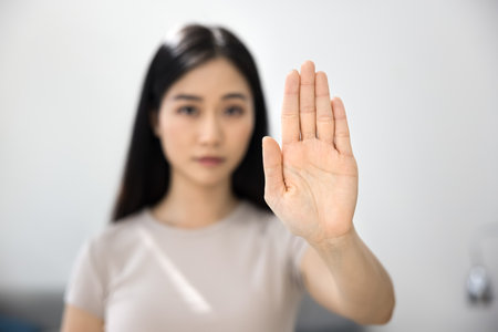 Young 30s Asian woman with serious expression extends her palm toward camera, making stop or refusal gesture. Rejection, protect personal boundaries, or protest in personal or social context, close upの写真素材