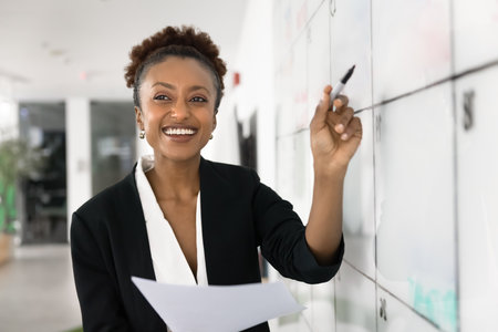 Strategic mindset and creative focus. Smiling young Black woman task manager strategist stand near glass timeline board hold paper report reflect with marker before mapping goals on planning calendarの写真素材
