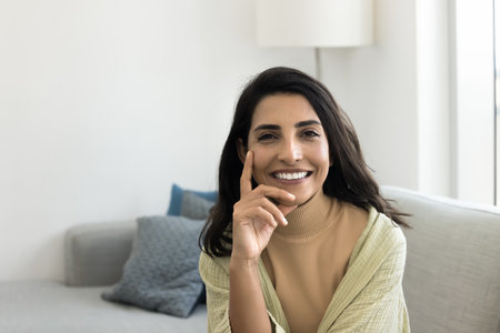 Head shot of beautiful Brazilian young 30s woman dressed in elegant, casual clothes staring at camera seated alone on comfortable couch in modern living room at own or rented apartment, home portraitの写真素材