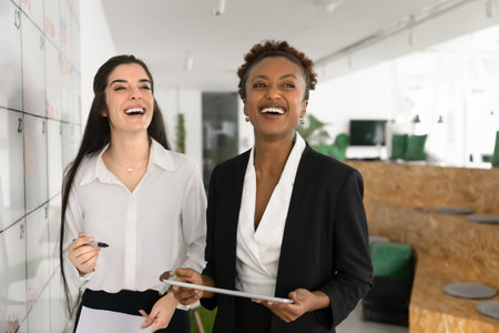 Collaborative planning with laughter. Joyful laughing multicultural team of two young women coworkers having fun tracking tasks progress syncing project data between digital pad and visual task boardの写真素材