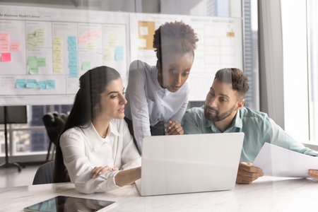 Project development synergy. Young multiethnic businesspeople gather at desk around notebook in modern office to brainstorm marketing solutions review sales reports discuss research in Agile workspaceの写真素材