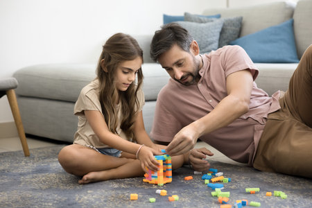 Middle-aged loving father playing colorful cubes with little daughter, settled down comfortably on warm floor, focused on creative activity, helps with construction, enjoy family quality time togetherの写真素材