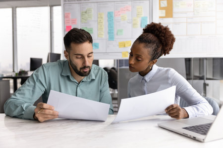 Collaborative paperwork. 2 diverse young business colleagues project managers planners review documents at workplace analyze finance reports discuss workflow innovations at desk near Agile task boardの写真素材