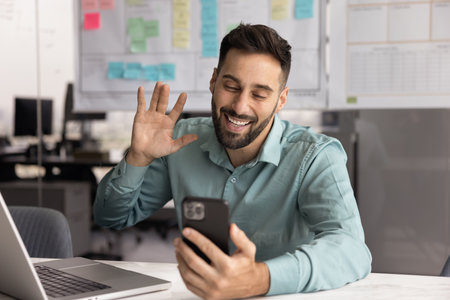 Virtual communication. Happy young male manager office employee distracted from office work on notebook to make answer video call on phone wave hand greet colleague client on meeting in conference appの写真素材