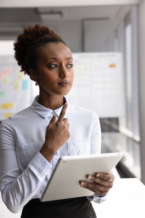 Solving problem using digital tools. Pensive young female entrepreneur project manager creative planner standing in office holding tablet pc looking aside making business decision considering strategyの写真素材