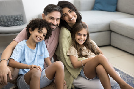 Happy multiethnic couple cuddling their cute little son and daughter seated on rug, expressing warmth, feeling unconditional love, enjoy moment of tenderness in living room. Time together, family bondの写真素材