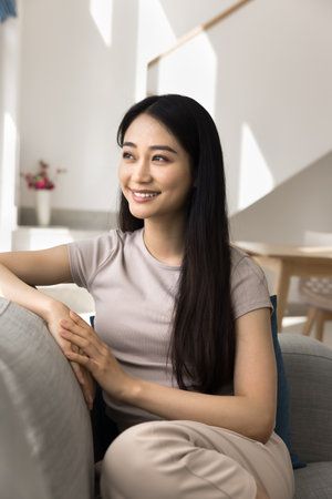 Serene beautiful woman in casual clothes sits comfortable on cozy couch, looks into distance with contented smile, spend carefree leisure in contemporary sunlit living room alone at home. Restの写真素材