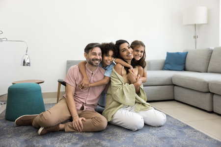 Loving multiethnic couple with little children sitting on rug in contemporary living room, laughing, looking away, enjoying time together on carefree weekend. Parenthood, homeowners family portraitの写真素材