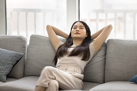 Peaceful young 25s Asian woman enjoying quiet moment on comfortable grey sofa, leaned back with...
