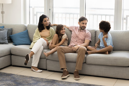 Multiethnic mother and father sitting on cozy couch with two school age 7s children, smiling, engaged in pleasant, trustworthy conversation. Family communication, active parenting, weekend togetherの写真素材