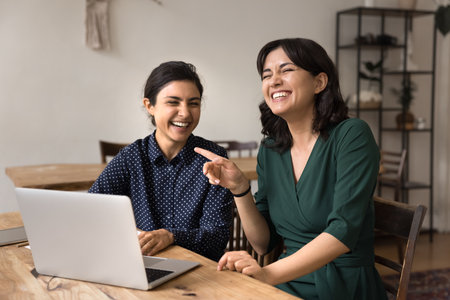 Joyful teamwork and friendly collaboration. Laughing overjoyed young businesswomen having fun at office desk develop startup on laptop discuss business idea with joy show energetic creative atmosphereの写真素材