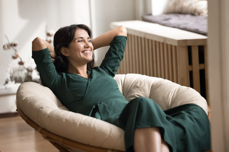 Cozy home relax. Calm smiling millennial woman recline in comfortable rattan armchair with pillow hands behind head enjoy lazy weekend in quiet living room personal comfort stress free moment indoorsの写真素材