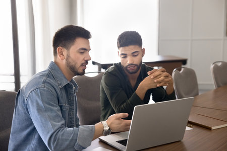 Manager presenting information on laptop to client sitting at table in modern office. Two colleagues...