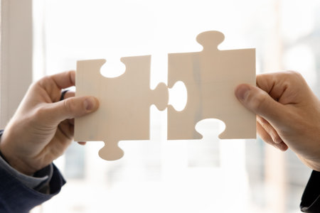 Two business colleagues joining jigsaw parts, holding pieces of puzzle, finding best successful solution together, starting collaboration, teamwork, partnership. Cropped shot of handsの写真素材