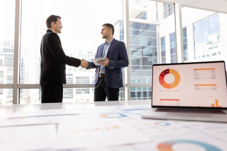 Positive male business partners closing deal in office, shaking hands behind table with marketing research results, project paper financial reports, finishing meeting with handshakeの写真素材