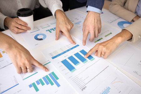 Hands of four analysts pointing at paper diagrams on meeting table high angle. Team of experts...