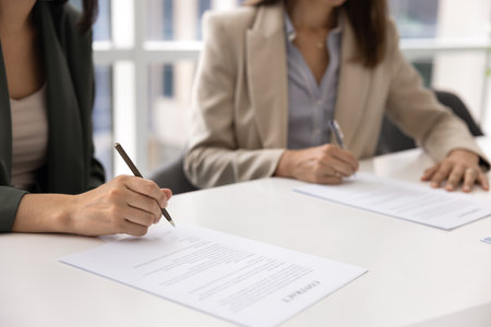 Two businesswomen getting agreement after negotiation meeting, putting signatures to paper document, signing contract, meeting at large office white table, holding pen, writing. Cropped shotの写真素材