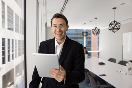 Happy handsome young business professional man in glasses and formalwear using tablet computer in office meeting room, standing at large window, posing for portrait, smilingの写真素材