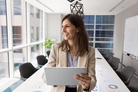 Cheerful successful company leader woman analyzing KPI, benchmarks of growth on tablet, monitoring internet product promotion, holding digital gadget, looking away, smiling, thinkingの写真素材