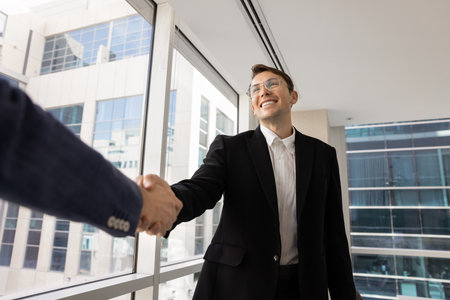 Happy ambitious young executive man in classy jacket shaking hands with male partner on meeting,...