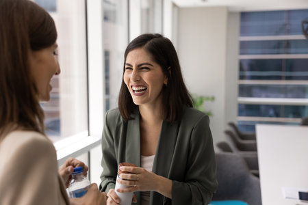 Cheerful young Latin business professional chatting and laughing with female colleague on work break, talking to colleague, having fun with office friend, taking break for hot drink, holding thermosの写真素材