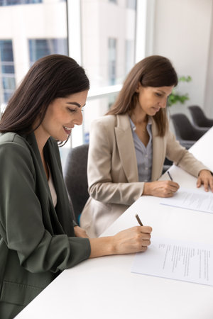 Two young businesswomen putting signature to contract on negotiation, meeting at large table,...