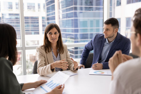 Positive Caucasian businesswoman talking to coworkers at meeting table, sharing ideas for brainstorming with team. Business group discussing teamwork, project marketing reportsの写真素材