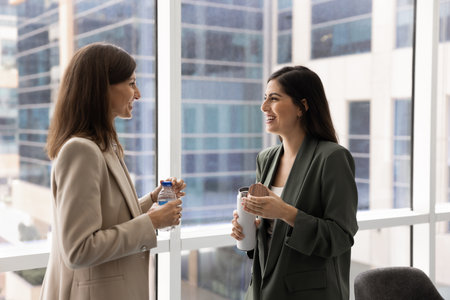 Two happy diverse female coworkers talking in office hall on coffee break, holding bottles with hot drink, water, standing at large window, chatting, laughing, enjoying leisure away from workplaceの写真素材