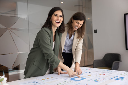Two cheerful young female business colleagues analyzing product development marketing data, discussing paper reports on large table, smiling, laughing, talking, meeting for teamworkの写真素材