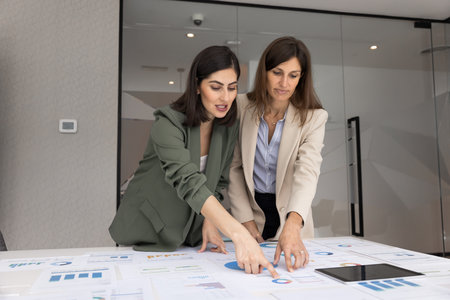 Two diverse female analysts working on project marketing strategy together, analyzing research results, talking at table with paper financial sales reports, pointing at diagramsの写真素材