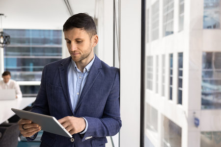 Serious successful Caucasian business executive man using online web professional service on tablet computer, standing at window in office space with colleague working in backgroundの写真素材