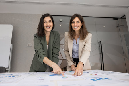 Two positive young product manager women working together at meeting table with marketing sales paper reports, analyzing charts, looking at camera, smiling, laughing, posing for portraitの写真素材