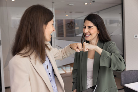 Cheerful young female business partners giving fist bump, smiling, laughing, discussing successful project, job success, achievement, sales growth, high marketing benchmarksの写真素材