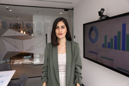 Confident beautiful young Latin business woman posing in office presentation room with electronic...