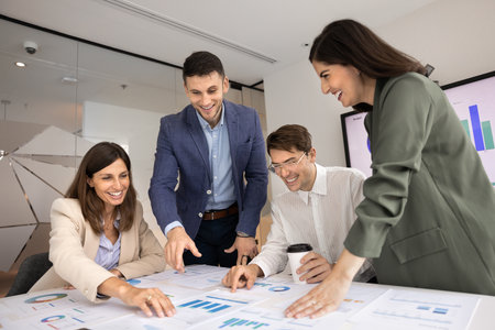 Team of four happy agency coworkers analyzing paper marketing reports on meeting table, pointing at charts, smiling, laughing, discussing ideas for successful startup strategy, sales growthの写真素材