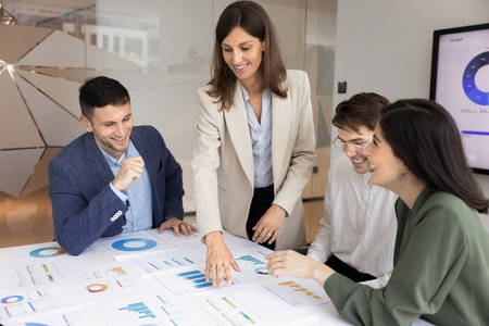 Business product manager woman presenting sales reports to team of coworkers on corporate meeting, showing paper charts on table. Team of colleagues discussing marketing strategyの写真素材