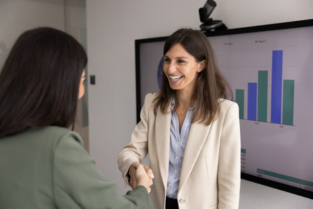 Happy female product manager woman shaking hands with female coworker at presentation board with marketing report. Business teacher giving handshake to trainee after training seminarの写真素材