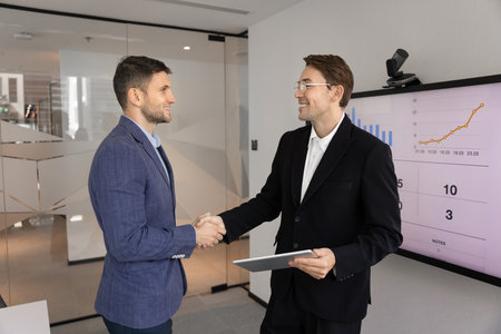 Positive young male professional coworkers shaking hands on presentation meeting, talking at electronic board, discussing collaboration. Businessman thanking coach for training seminar with handshakeの写真素材
