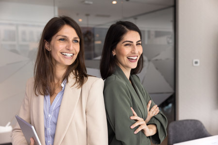 Two happy successful businesswomen posing for company profile portrait together, standing close together with shoulders touch, looking away, smiling, laughing, promoting women led businessの写真素材
