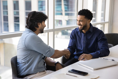 Successful partnership. Two happy friendly diverse businessmen shake hands at office desk making deal satisfied with effective collaboration. Grateful male intern appreciate mentor for help assistanceの写真素材